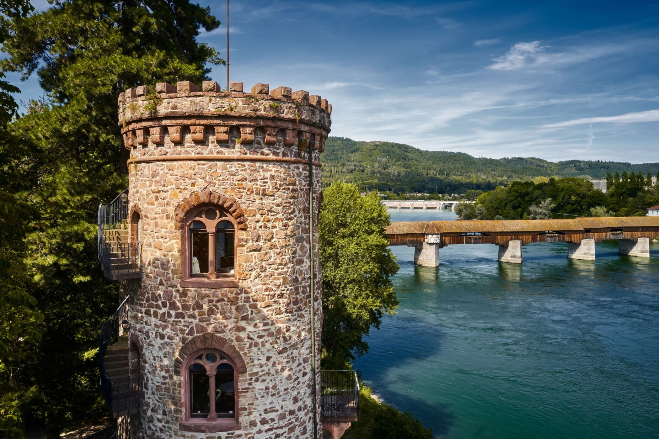 Steiniger Turm neben Fluss und Holzbrücke, umgeben von Bäumen und Hügeln.