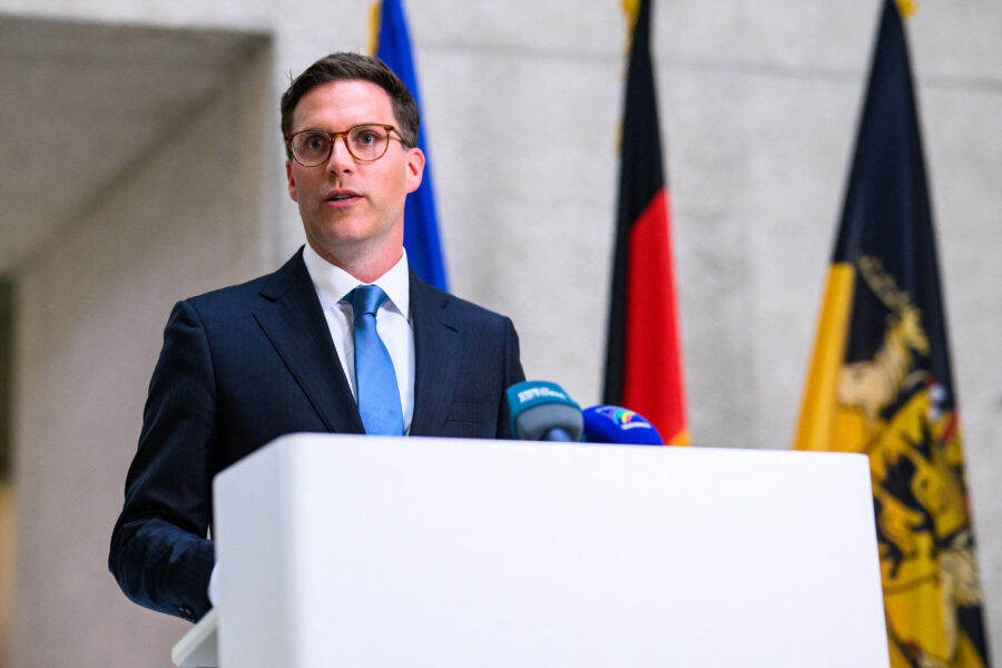 Mann im Anzug am Rednerpult mit Mikrofonen vor Flaggen im Hintergrund.