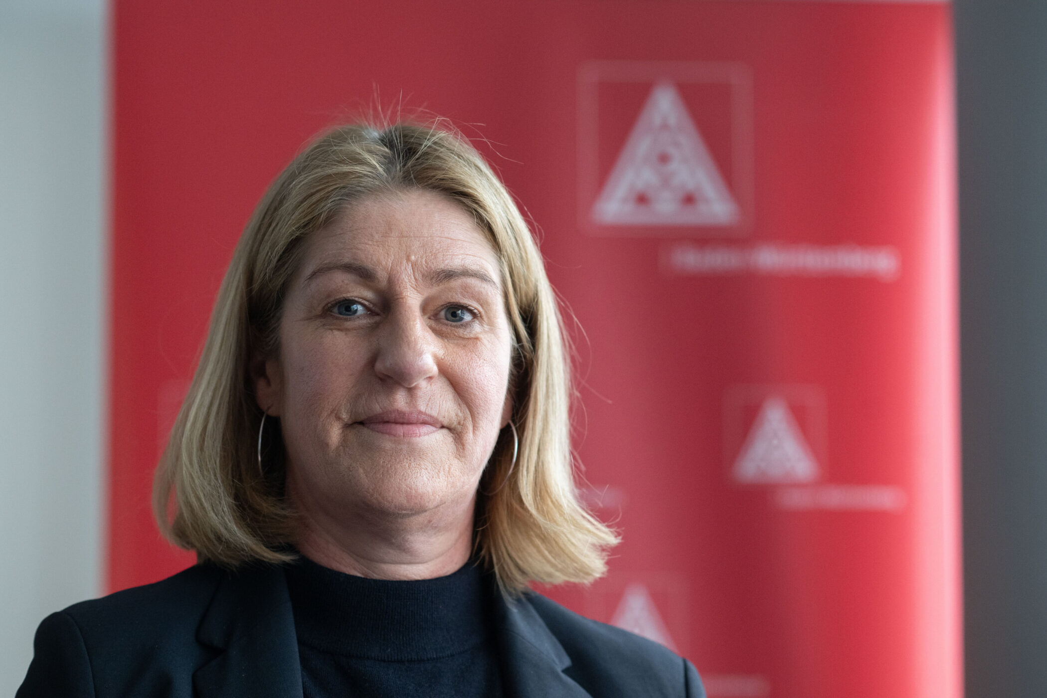 Frau mit blonden Haaren vor rotem Hintergrund mit Dreieckslogo und Text.