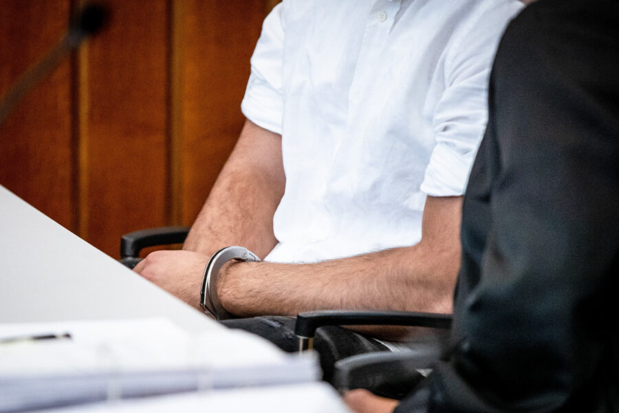 Mann in weißem Hemd sitzt, Hände gefesselt. Rechteckiger Tisch im Vordergrund.