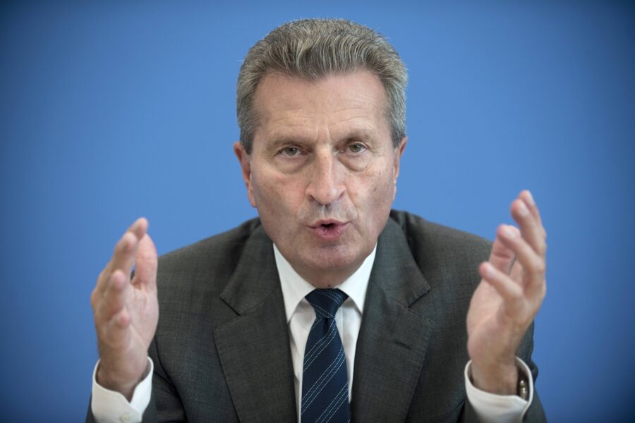 Älterer Mann im Anzug spricht mit erhobenen Händen vor blauem Hintergrund.