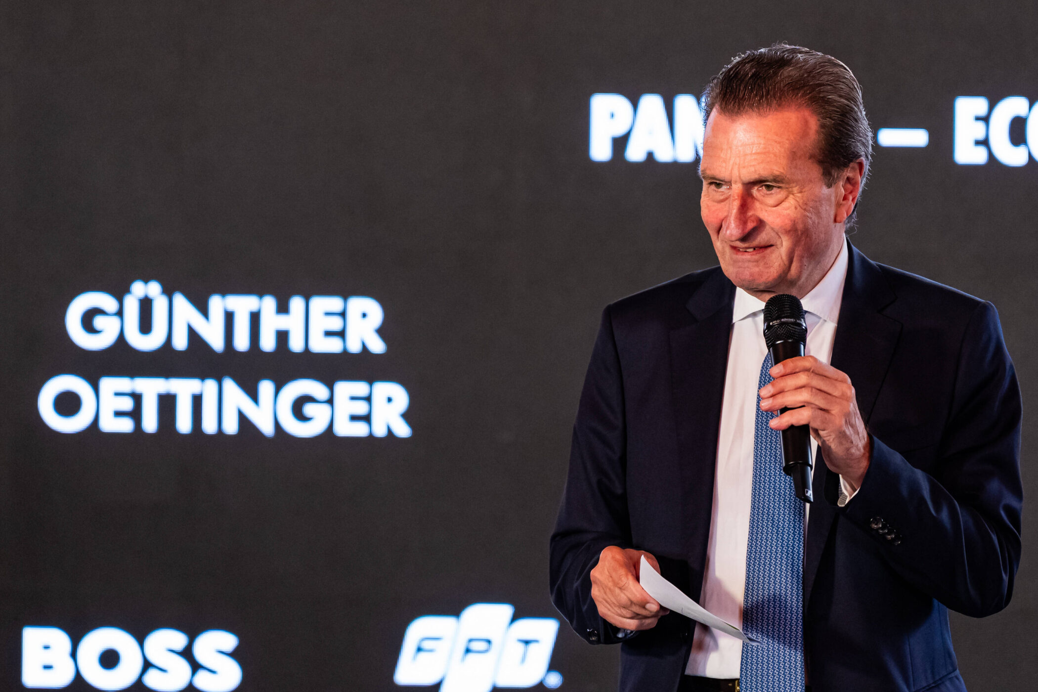 Mann im Anzug hält Mikrofon, Text "Günther Oettinger" auf schwarzem Hintergrund.