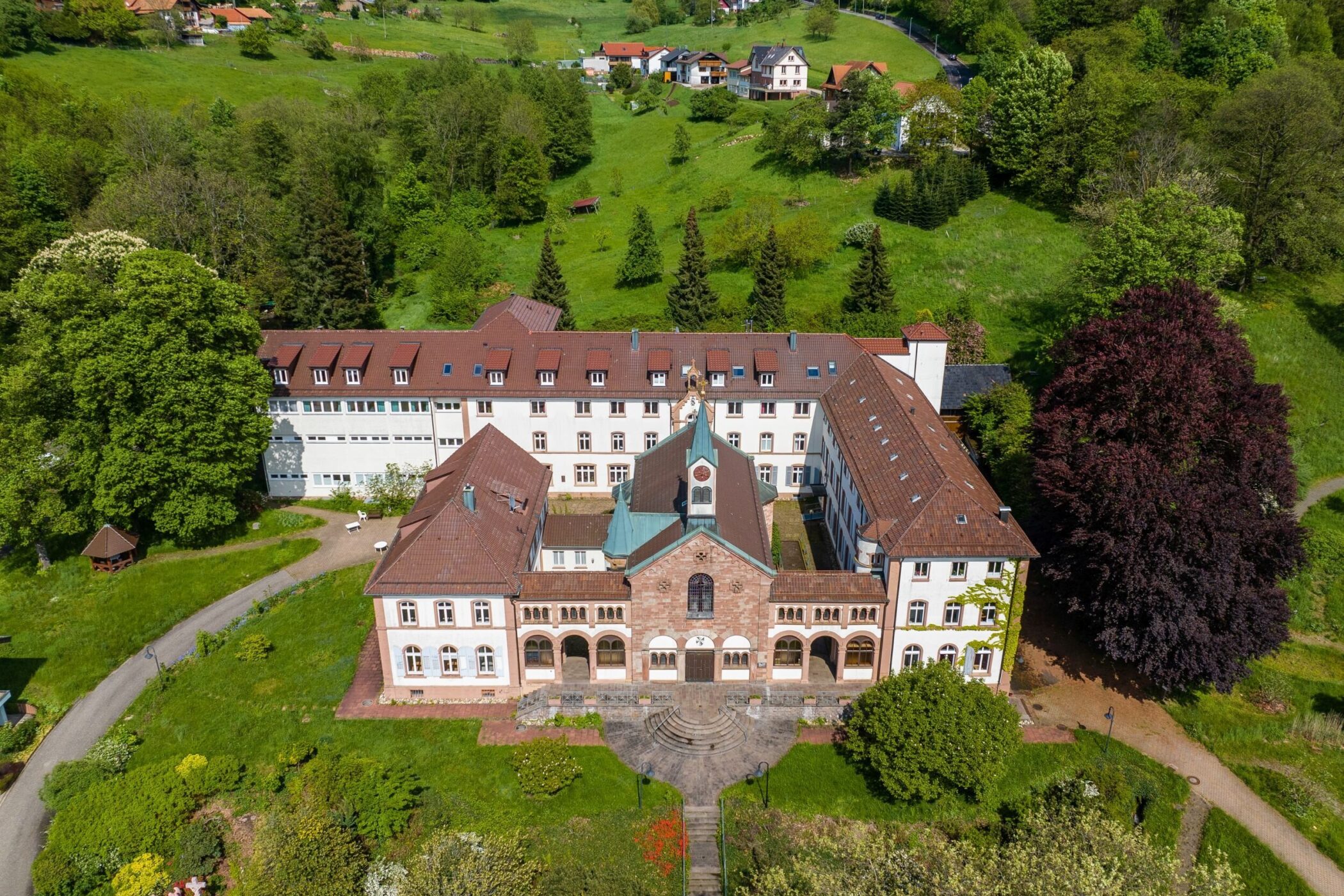 Großes Gebäude mit roten Dächern, umgeben von grüner Landschaft und Bäumen.