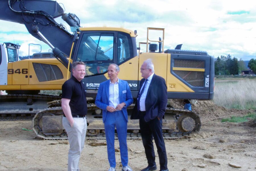 Drei Männer stehen vor einem gelben Bagger auf einer Baustelle.