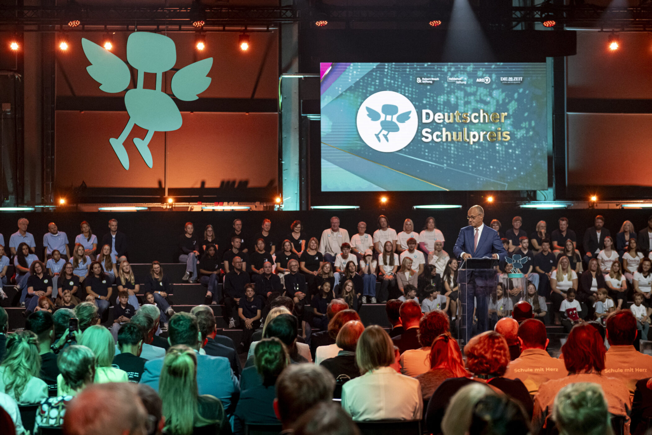 Person spricht auf Bühne vor Publikum, Logo und Text "Deutscher Schulpreis" auf Bildschirm.