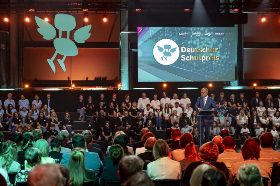 Person spricht auf Bühne vor Publikum, Logo und Text "Deutscher Schulpreis" auf Bildschirm.