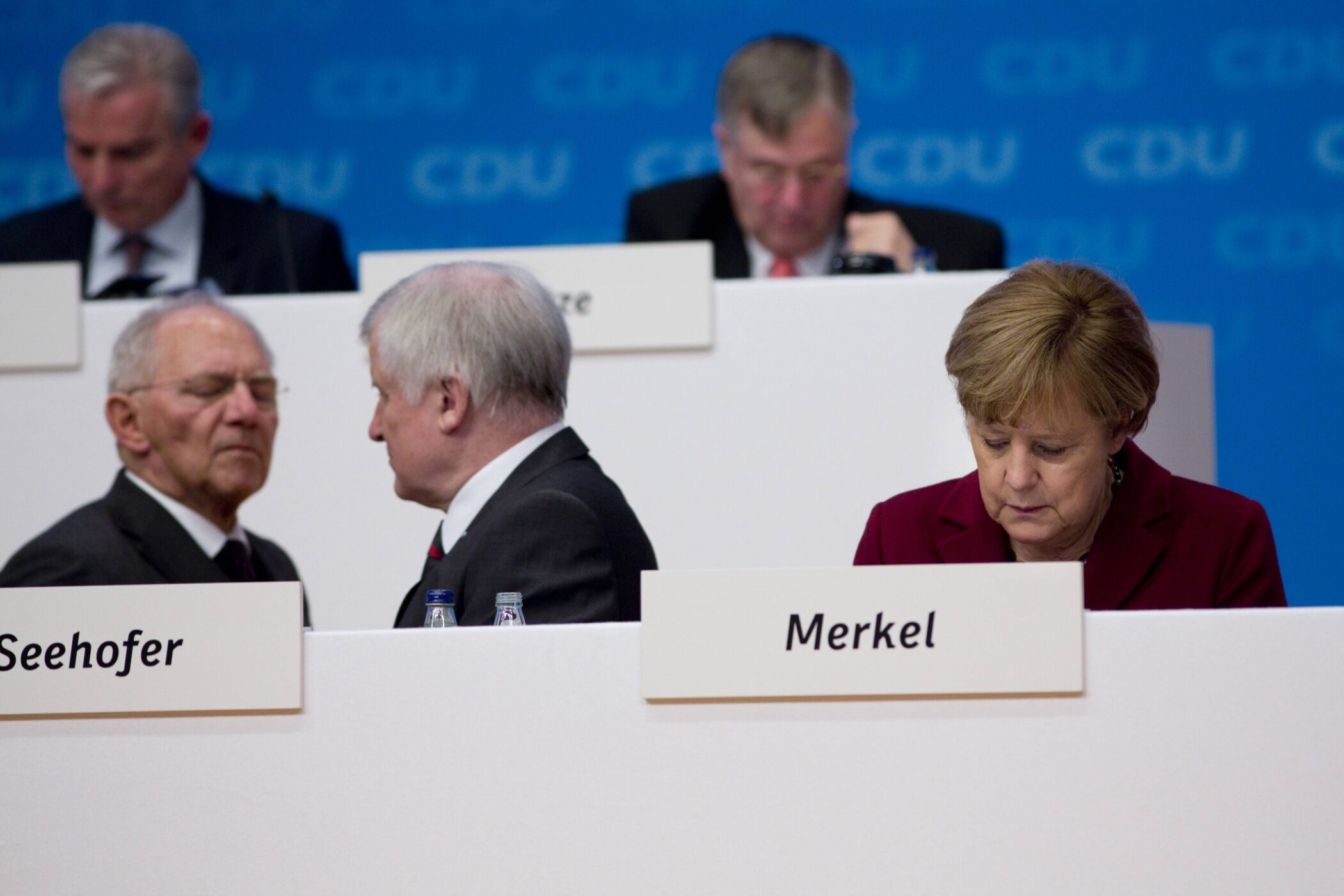 Fünf Personen bei Konferenz, Namensschilder Seehofer und Merkel sichtbar.