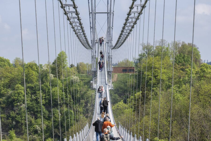 Menschen auf Hängebrücke über Wald. Himmel teils bewölkt.
