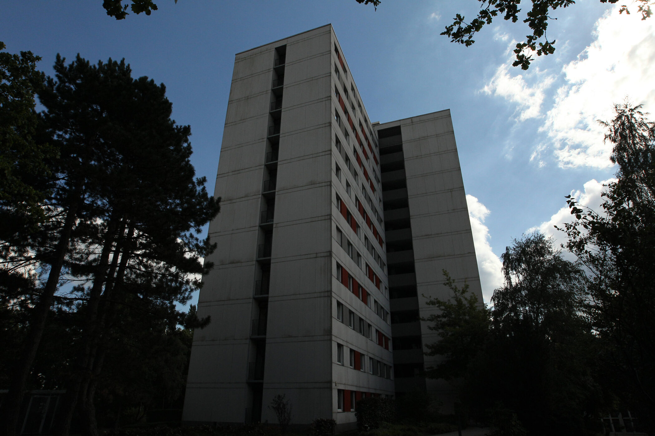 Hohes, rechteckiges Gebäude mit roten und weißen Fensterläden, Bäume im Vordergrund.