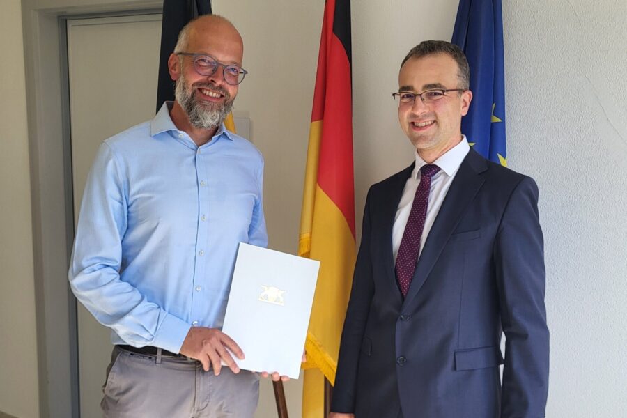Zwei Männer im Anzug, Mann links hält Dokument, deutsche und EU-Flagge im Hintergrund.