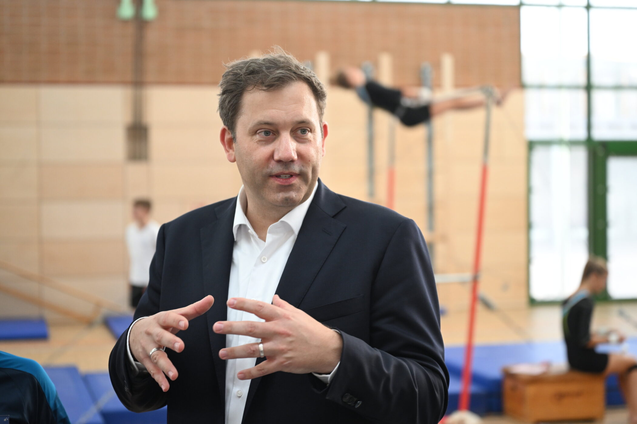 Ein Mann im Anzug gestikuliert in einer Turnhalle. Im Hintergrund turnen Personen.