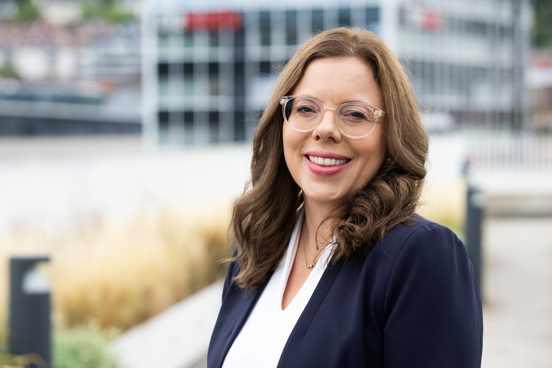 Eine Frau mit Brille lächelt vor einem unscharfen Bürogebäude.