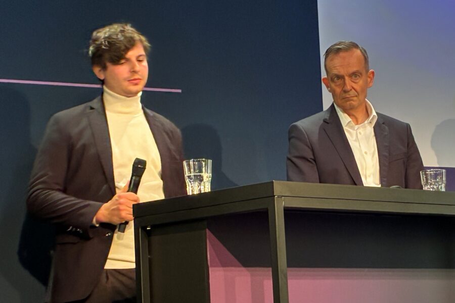 Zwei Männer in Anzügen stehen an einem Tisch mit Mikrofon und Wassergläsern.