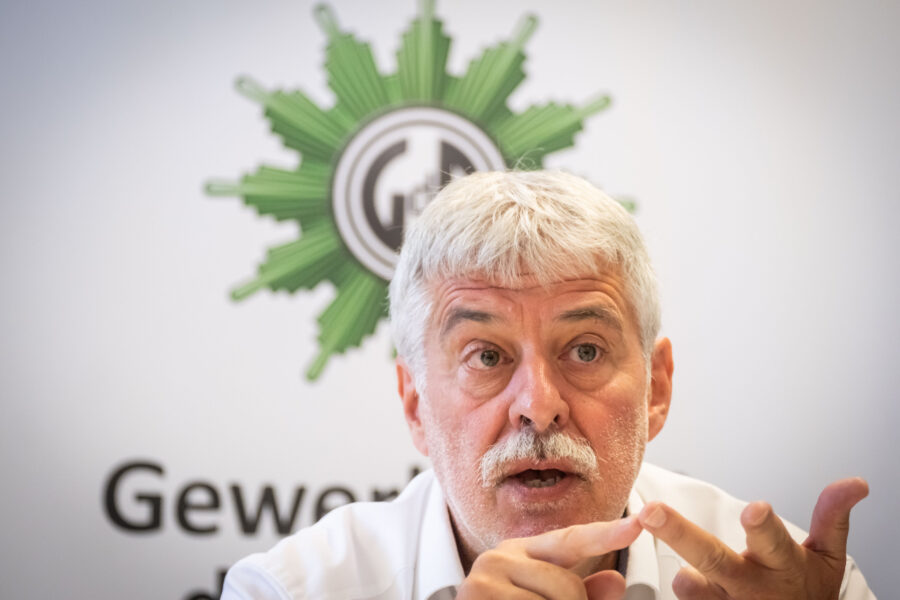 Älterer Mann, spricht und gestikuliert vor einem Polizeiemblem.