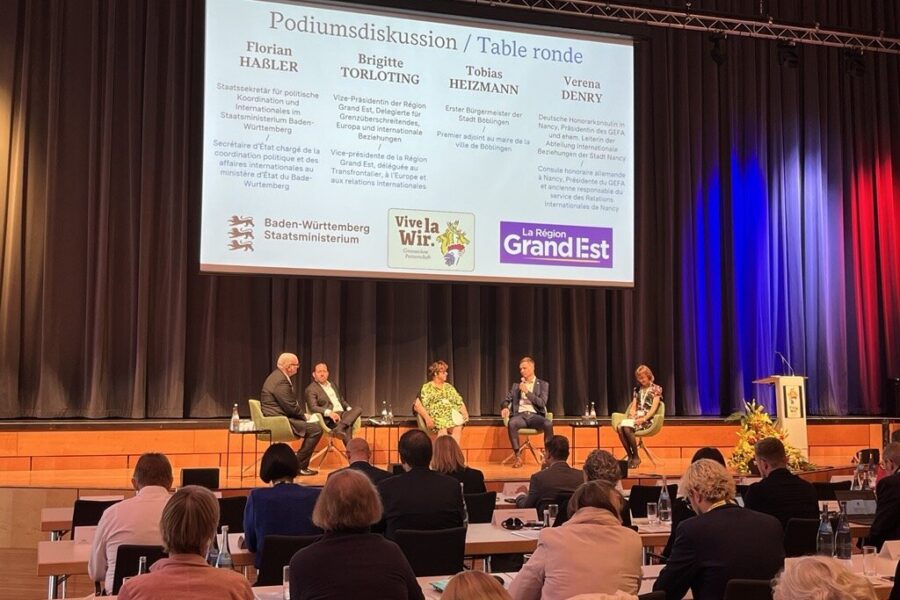Fünf Personen auf einer Bühne bei einer Podiumsdiskussion mit blau-rotem Vorhang.