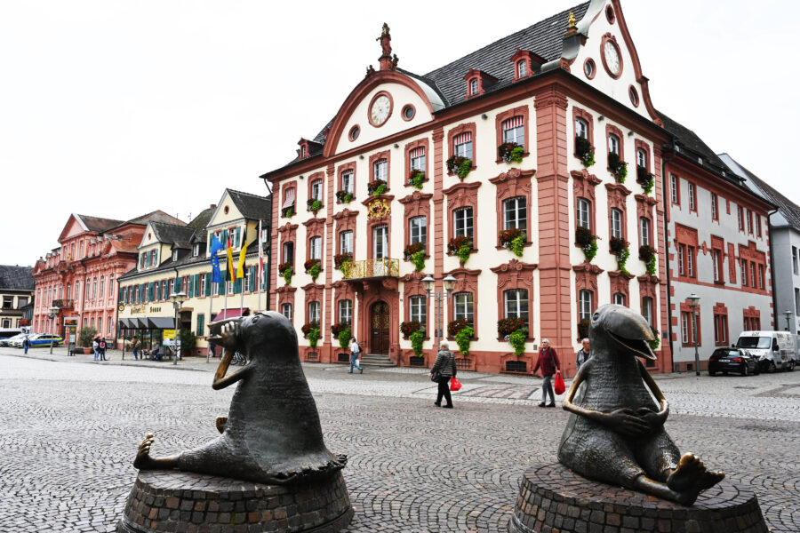 Rotes Gebäude mit Balkon, Blumen. Zwei bronzene Skulpturen, Personen auf Kopfsteinpflaster.