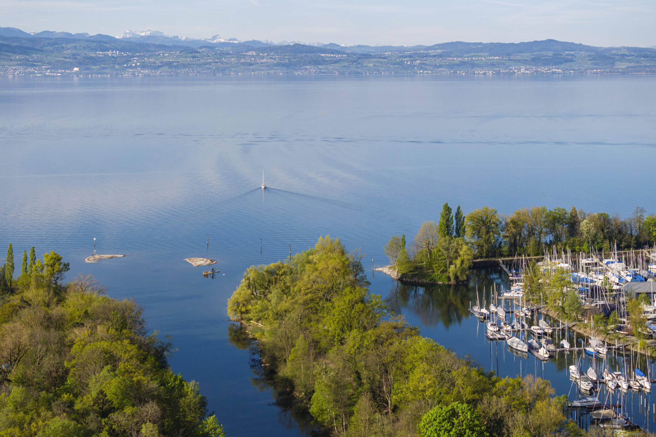Segelboot auf ruhigem See, grüne Inseln, Berge im Hintergrund, Yachthafen rechts.