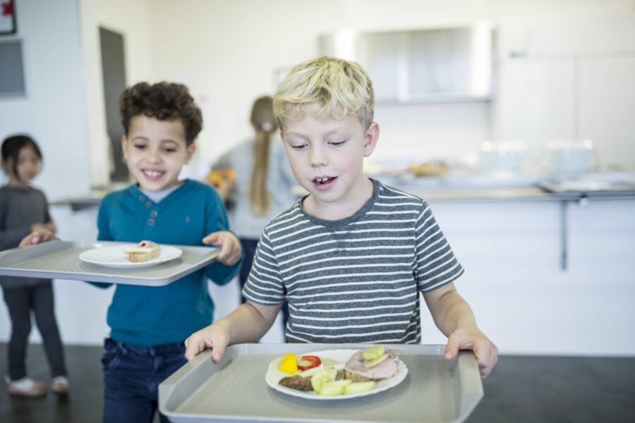 Zwei Kinder mit Tabletts und Tellern mit Essen in einem Speisesaal.