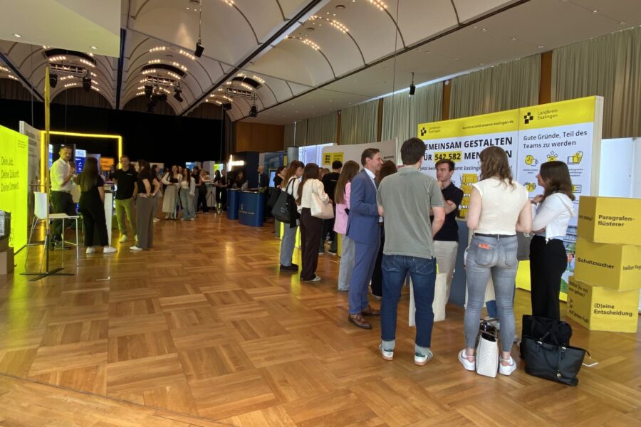 Menschen in einer Halle sprechen mit Ausstellern an gelben Infoständen.