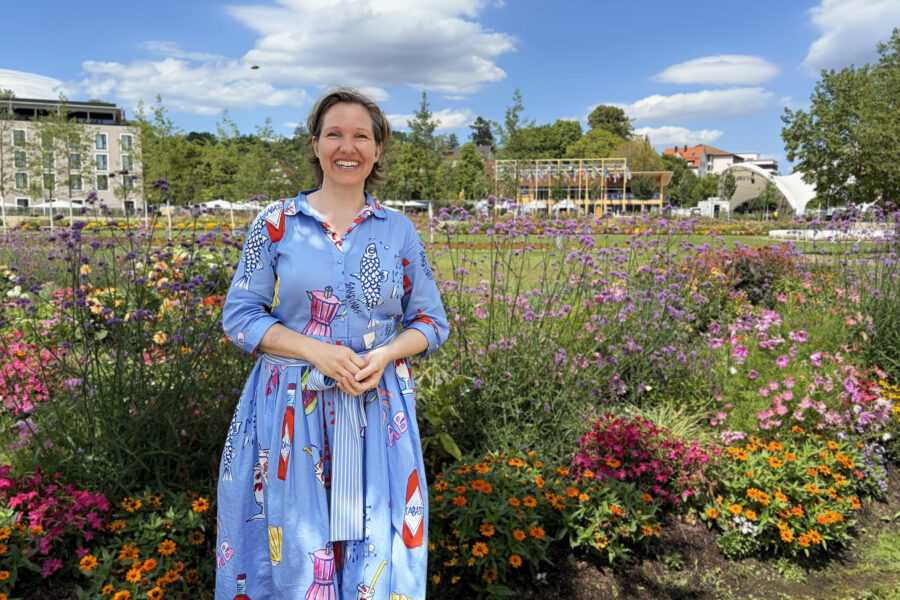 Person steht vor buntem Blumenbeet, Gebäude und blauer Himmel im Hintergrund.