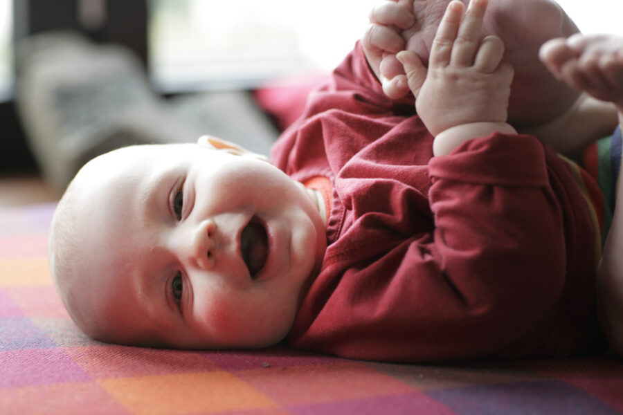Liegendendes Baby in rotem Shirt greift seine Füße auf bunt karierter Decke.