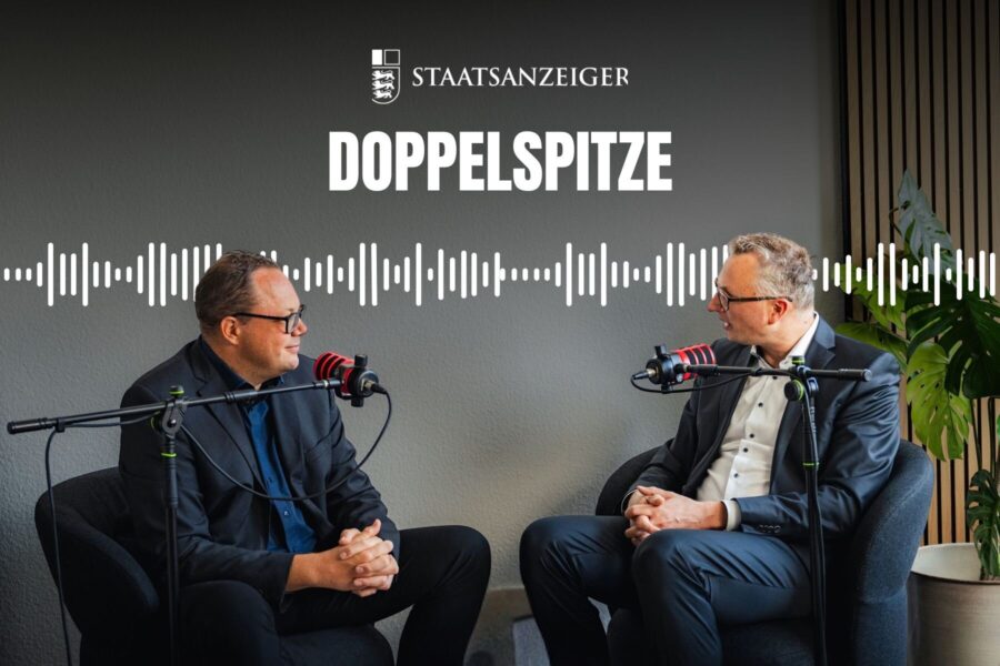 Zwei Männer in Sesseln sprechen in Mikrofone, oberhalb Schriftzug "STAATSANZEIGER DOPPELSPITZE".