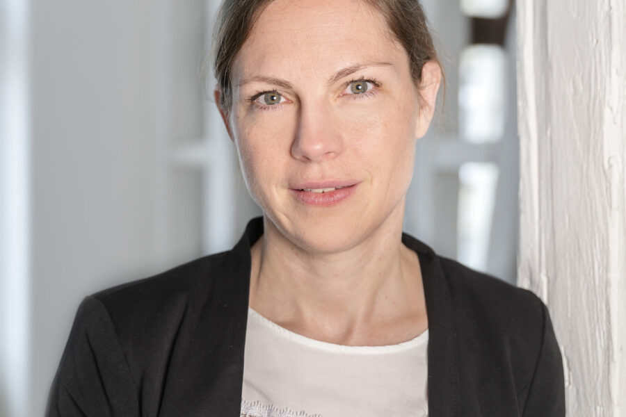 Frau mit dunklem Blazer und weißem Oberteil vor weißem Hintergrund.