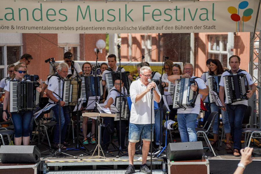 Gruppe mit Akkordeons auf einer Bühne, Mann singt am Mikrofon, Banner "Landes Musik Festival".