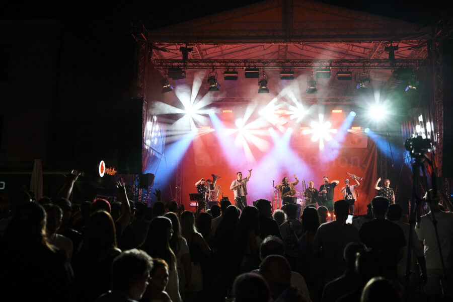 Bühne mit Band und Publikum im Vordergrund, Scheinwerfer in verschiedenen Farben.