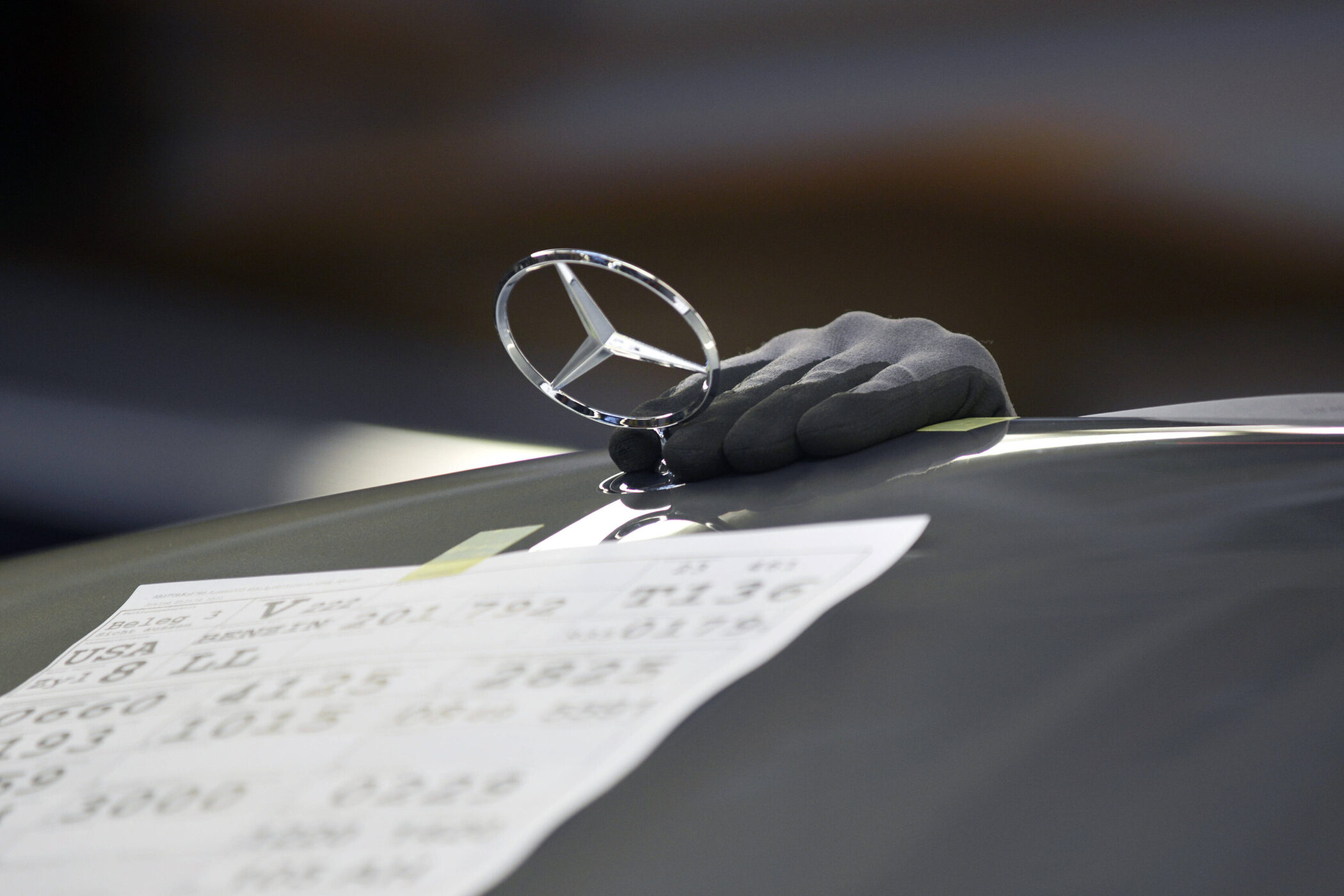 Hand mit Handschuh berührt Mercedes-Stern auf einem Auto, Papier im Vordergrund.