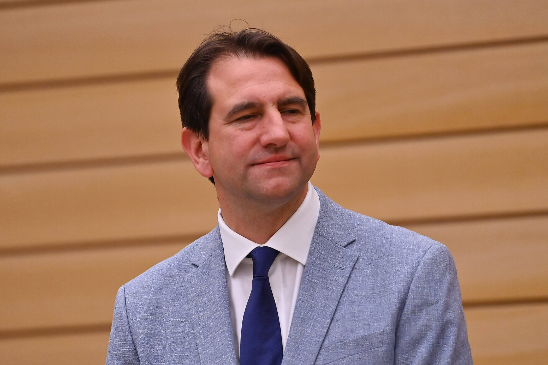 Mann in hellblauem Anzug und dunkler Krawatte vor holzgetäfelter Wand.