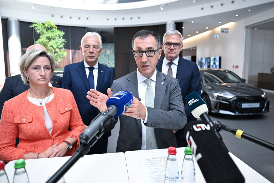 Fünf Personen stehen hinter Mikrofonen in einem Raum mit einem Auto im Hintergrund.