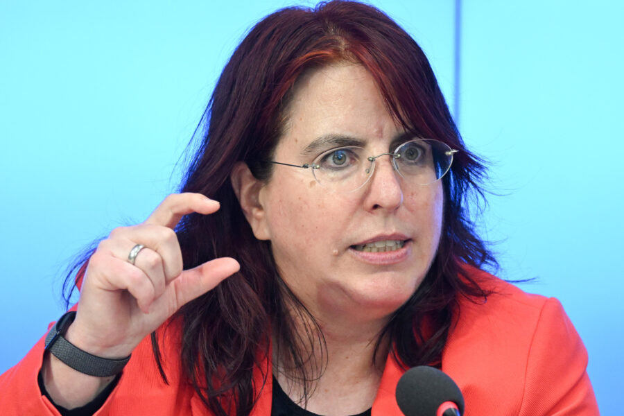 Frau mit Brille und rotem Blazer spricht, hebt eine Hand leicht an.