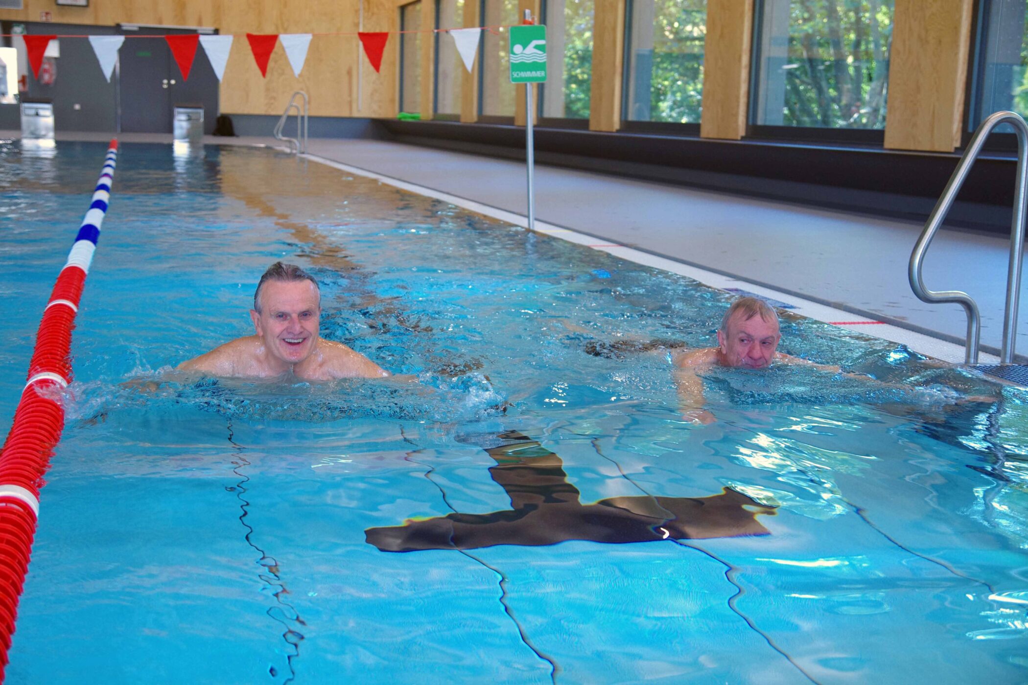 Zwei ältere Männer schwimmen in einem Hallenbad. Im Hintergrund rot-weiße Flaggen.