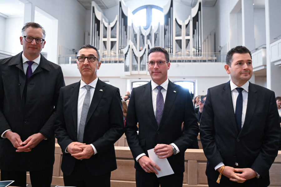 Vier Männer in Anzügen stehen nebeneinander, im Hintergrund eine Orgel.