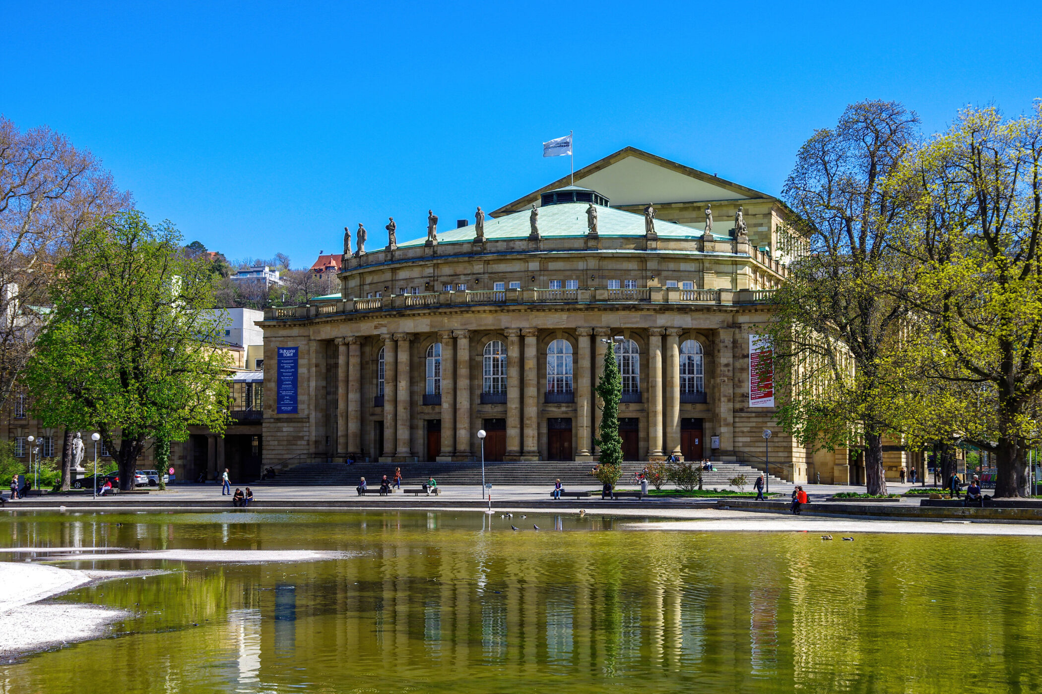 Rundes Gebäude mit Säulen und Dachfiguren, davor Teich und Bäume, Menschen auf Bänken.