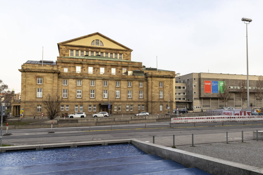Großes Gebäude mit gelber Fassade und vielen Fenstern, davor Straße und blaues Wasserbecken.