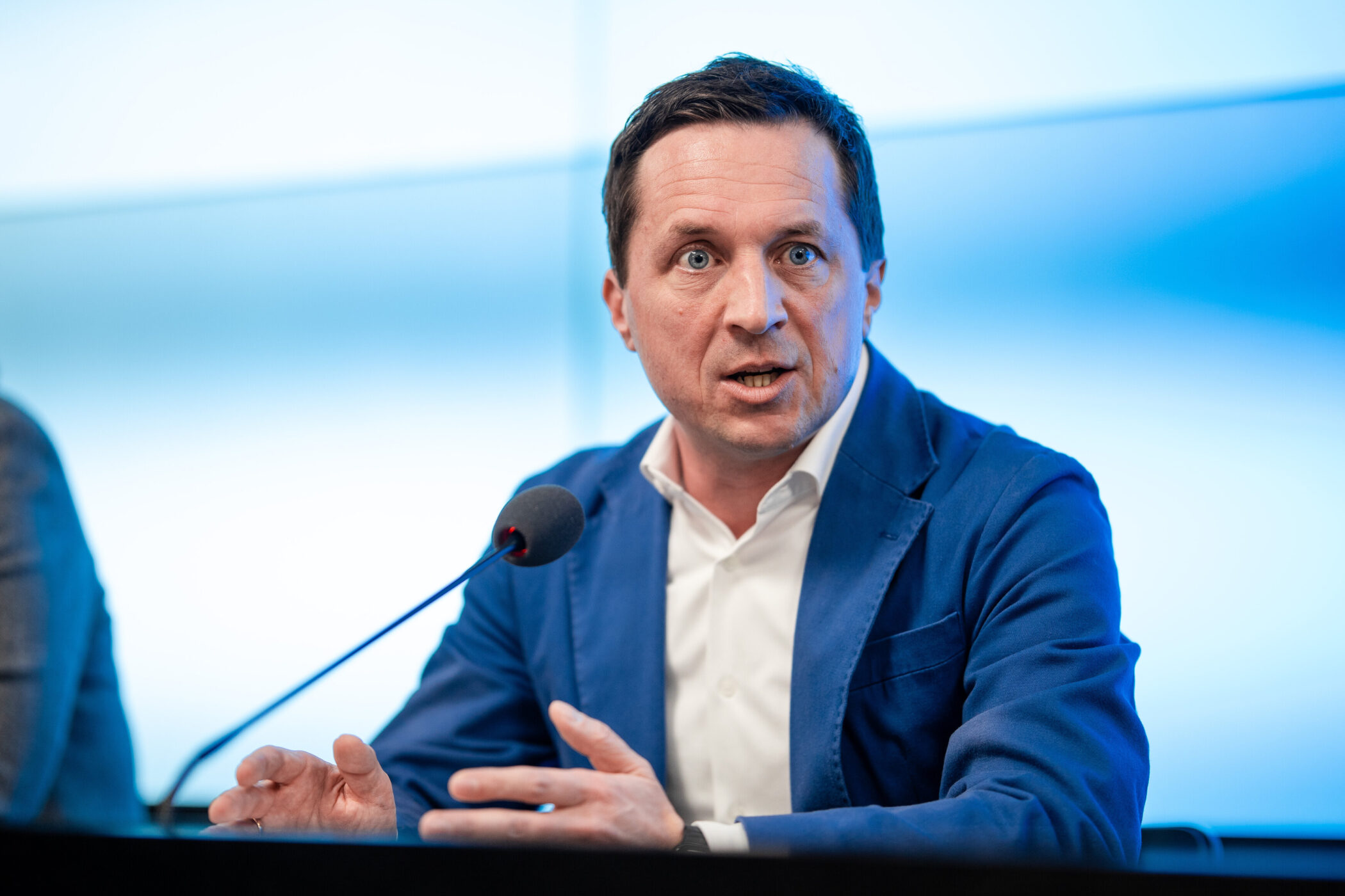Mann in blauem Anzug spricht in Mikrofon, blauer Hintergrund.