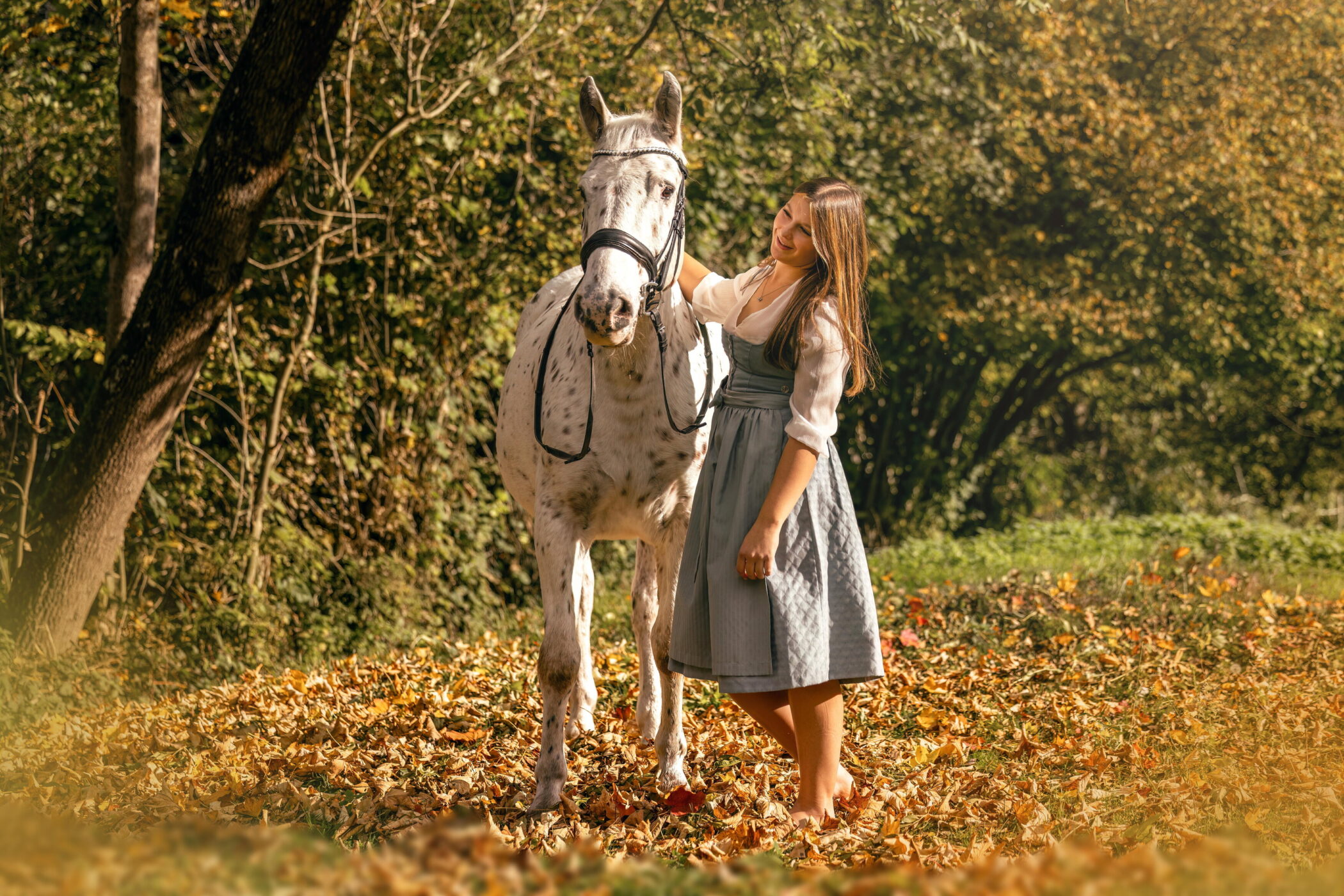 Frau im Dirndl steht neben einem gescheckten Pferd im herbstlichen Wald.