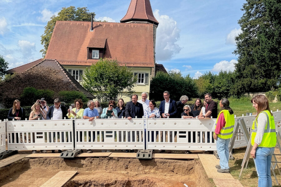 Gruppe von Menschen hinter Absperrung vor einem großen Gebäude mit rotem Dach.