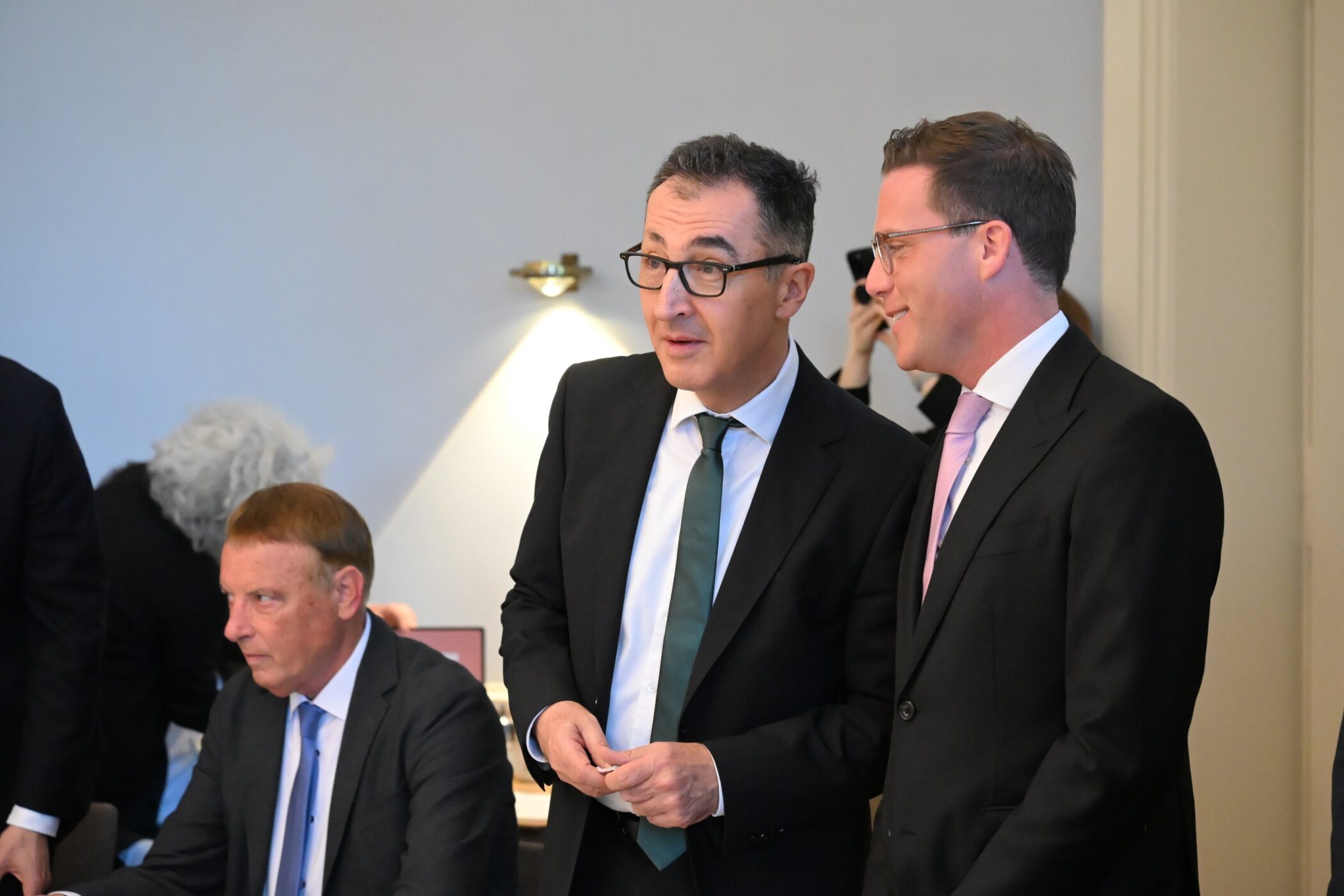 Drei Männer in Anzügen, zwei im Gespräch, einer sitzt.