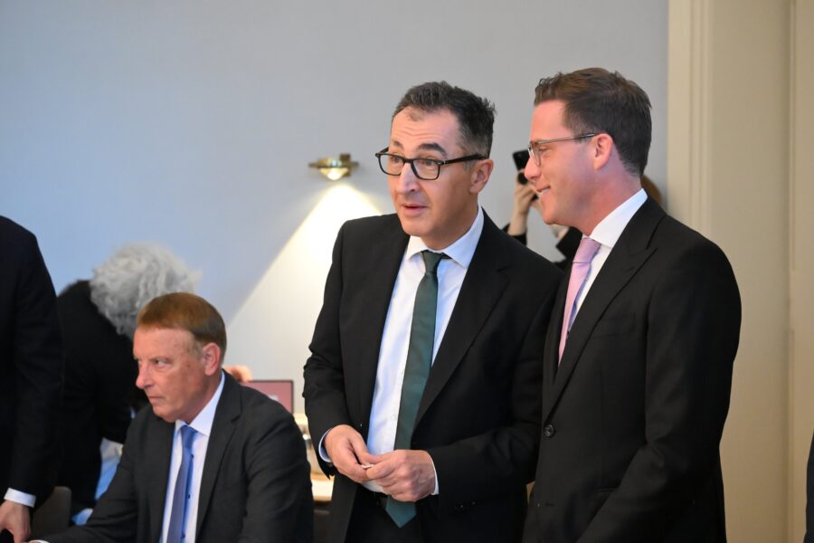 Drei Männer in Anzügen, zwei im Gespräch, einer sitzt.
