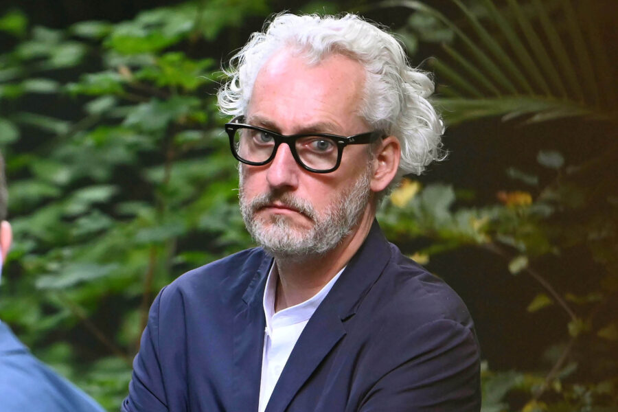 Person mit Brille und grauen Haaren blickt ernst, Hintergrund aus grünen Blättern.