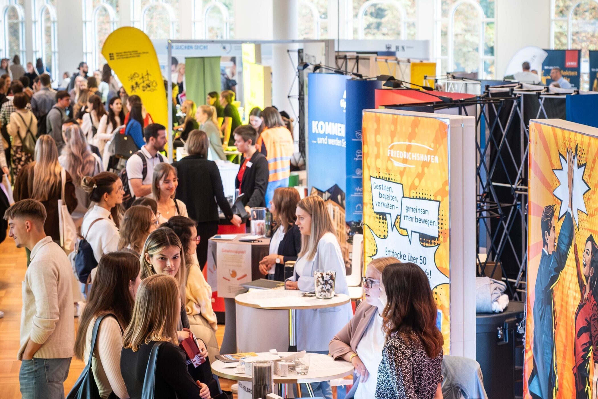Mehrere Menschen auf einer Messe mit bunten Informationsständen.