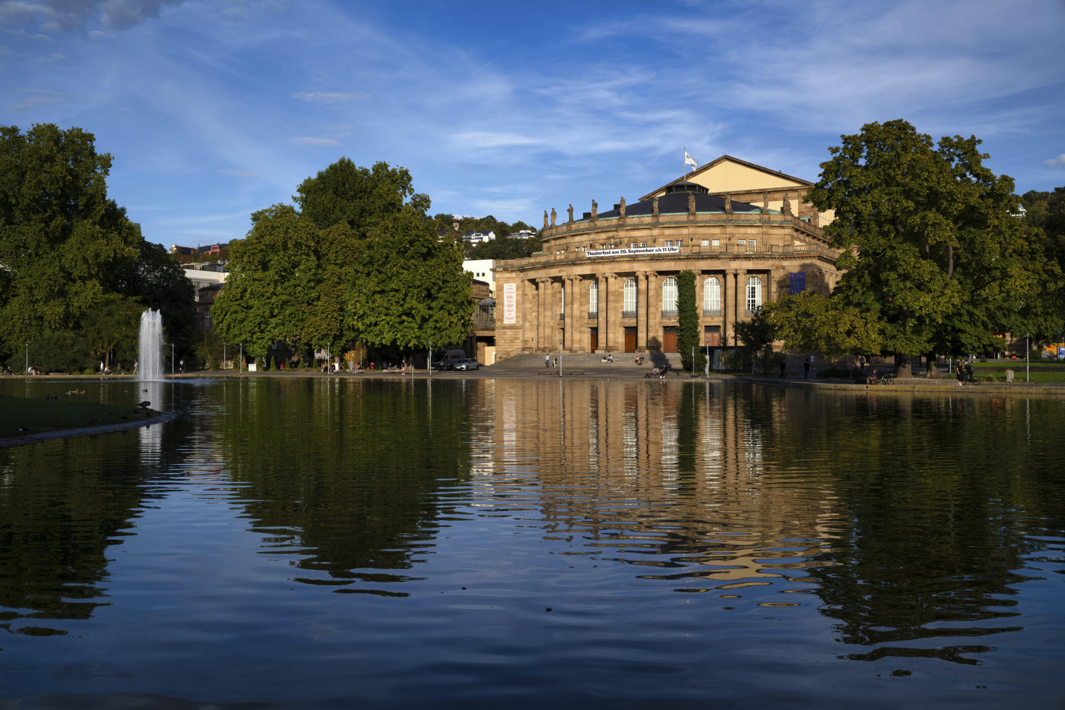 Gebäude mit Säulen hinter einem Teich, umgeben von Bäumen, blauer Himmel.