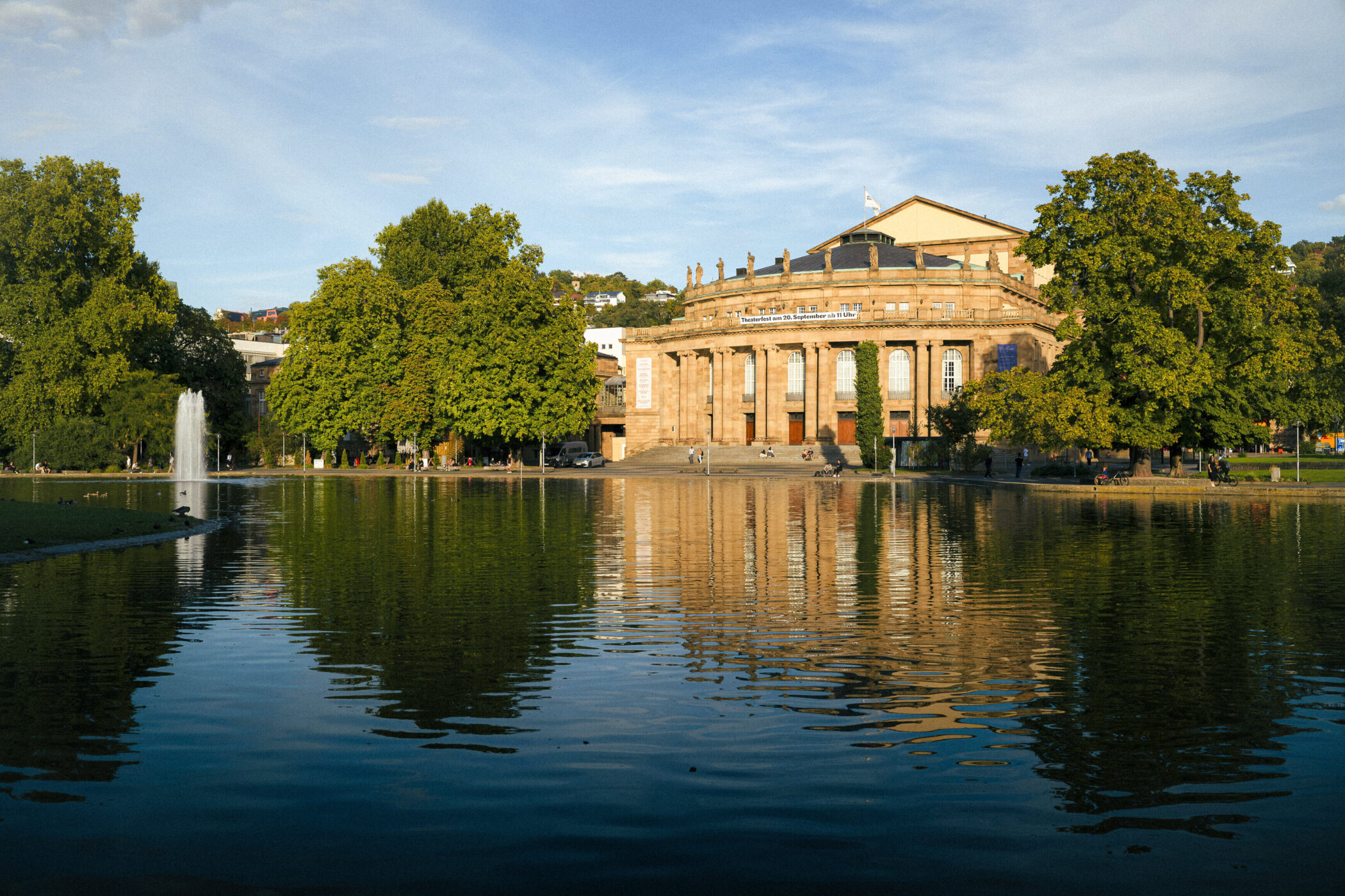 Großes Gebäude am See mit Bäumen und Wasserfontäne im Vordergrund.