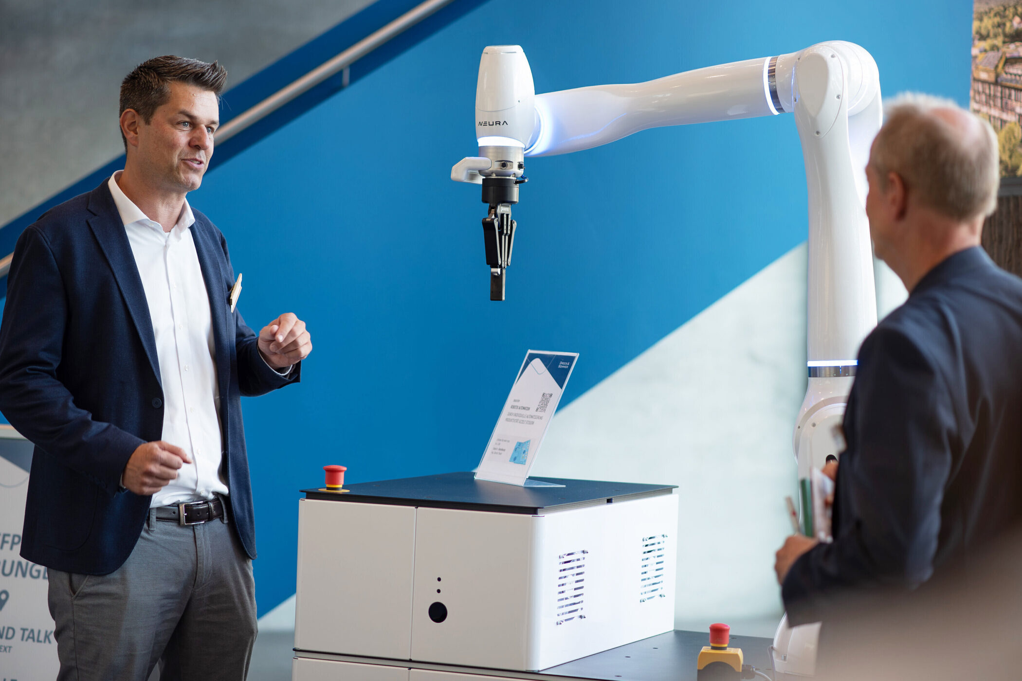 Zwei Männer stehen neben einem Roboterarm und einem weißen Gerät vor blauer Wand.