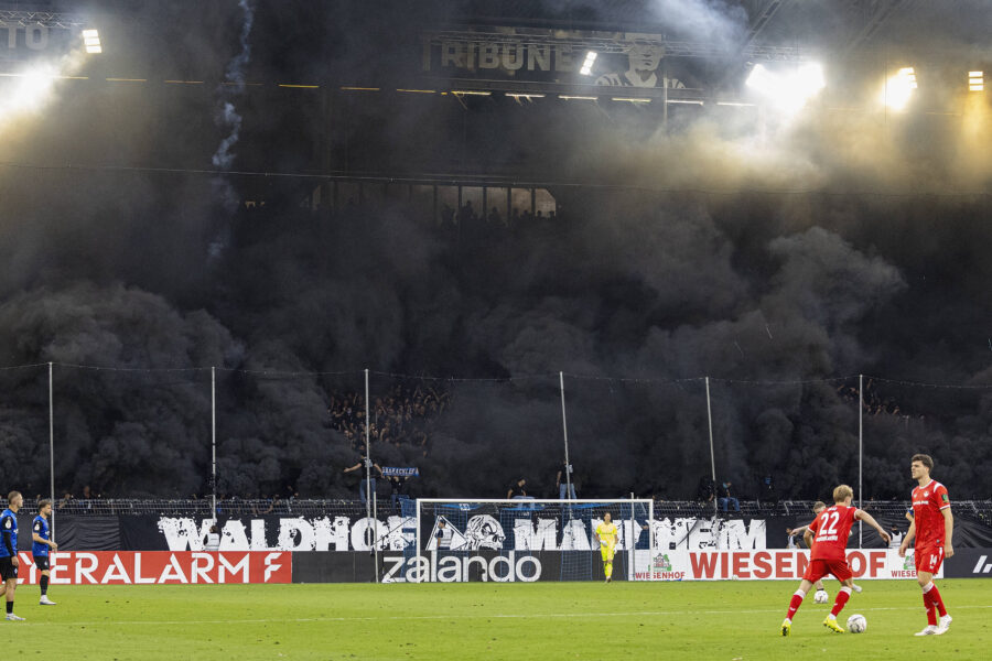 Zwei Fußballspieler in roten Trikots auf dem Spielfeld, dunkler Rauch im Hintergrund.