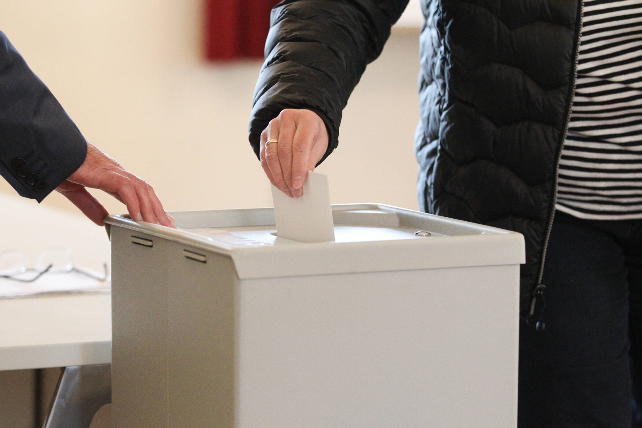 Zwei Personen werfen einen Zettel in eine graue Wahlurne ein.