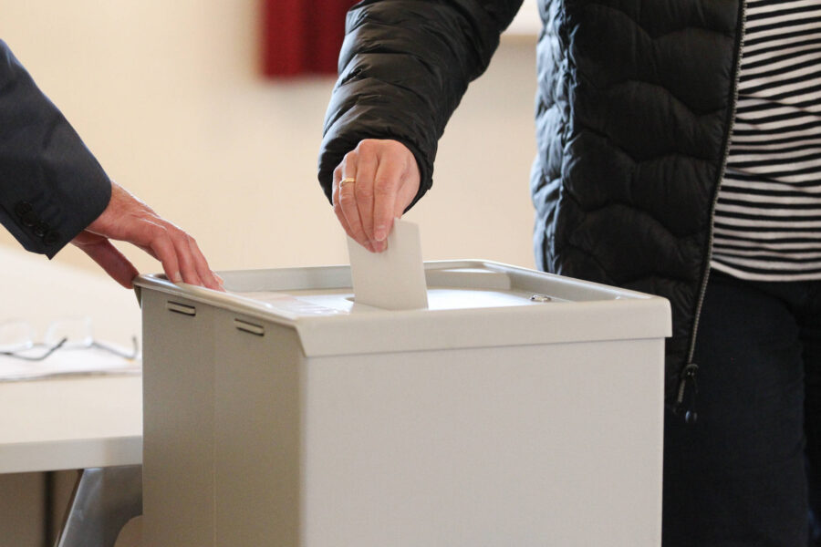 Zwei Personen werfen einen Zettel in eine graue Wahlurne ein.